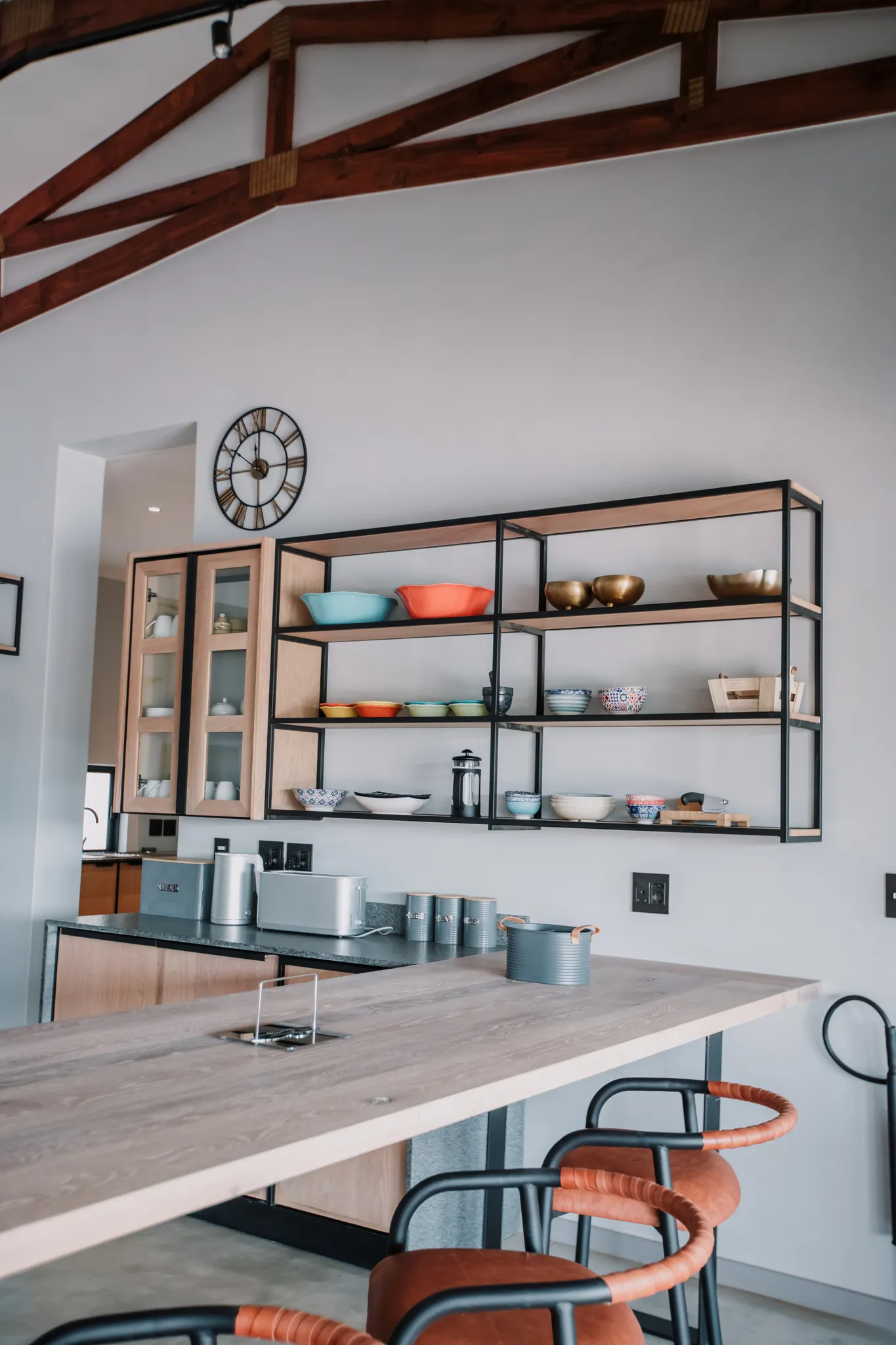 A kitchen with a wooden counter and barstools. Shelves with cutlery and utensils.